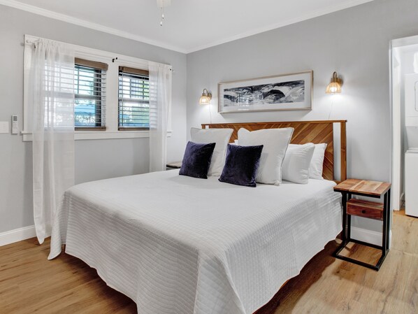 Ein ordentlich eingerichtetes Schlafzimmer mit einem großen Bett, zwei Nachttischen, einer Wandleuchte und einem gerahmten Bild über dem Bett.