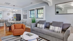 Una sala de estar moderna con un sofá gris, un sillón naranja y una mesa de centro blanca. Se aprecia un comedor con una mesa redonda y sillas, y una cocina al fondo.