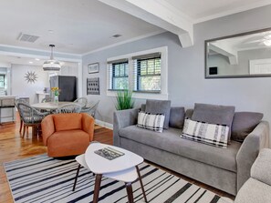 A modern living room with a grey sofa, orange armchair, and a white coffee table. There is a dining area with a round table and chairs, and a kitchen in the background.