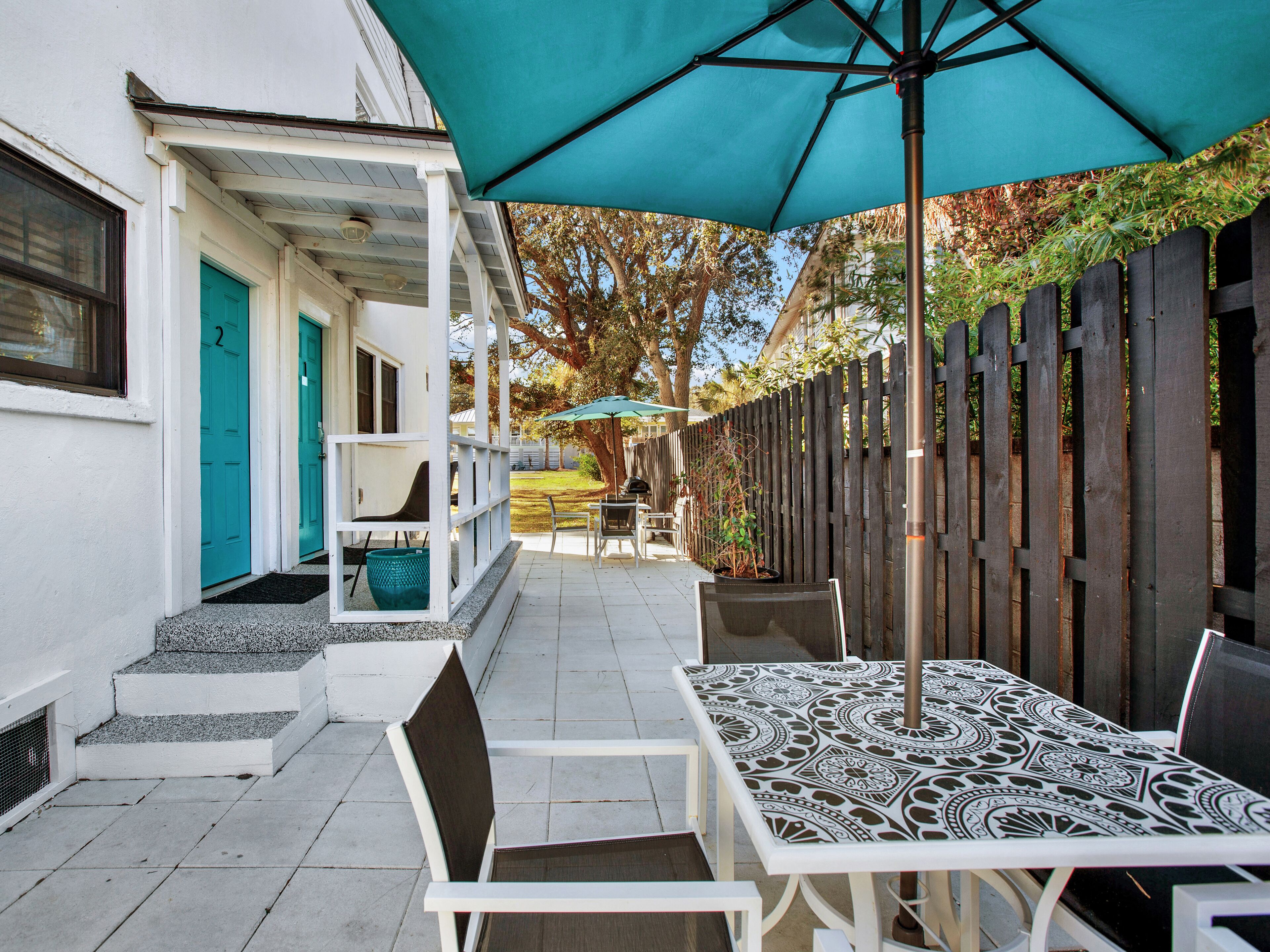 A patio with a table and chairs, a teal umbrella, and a wooden fence.