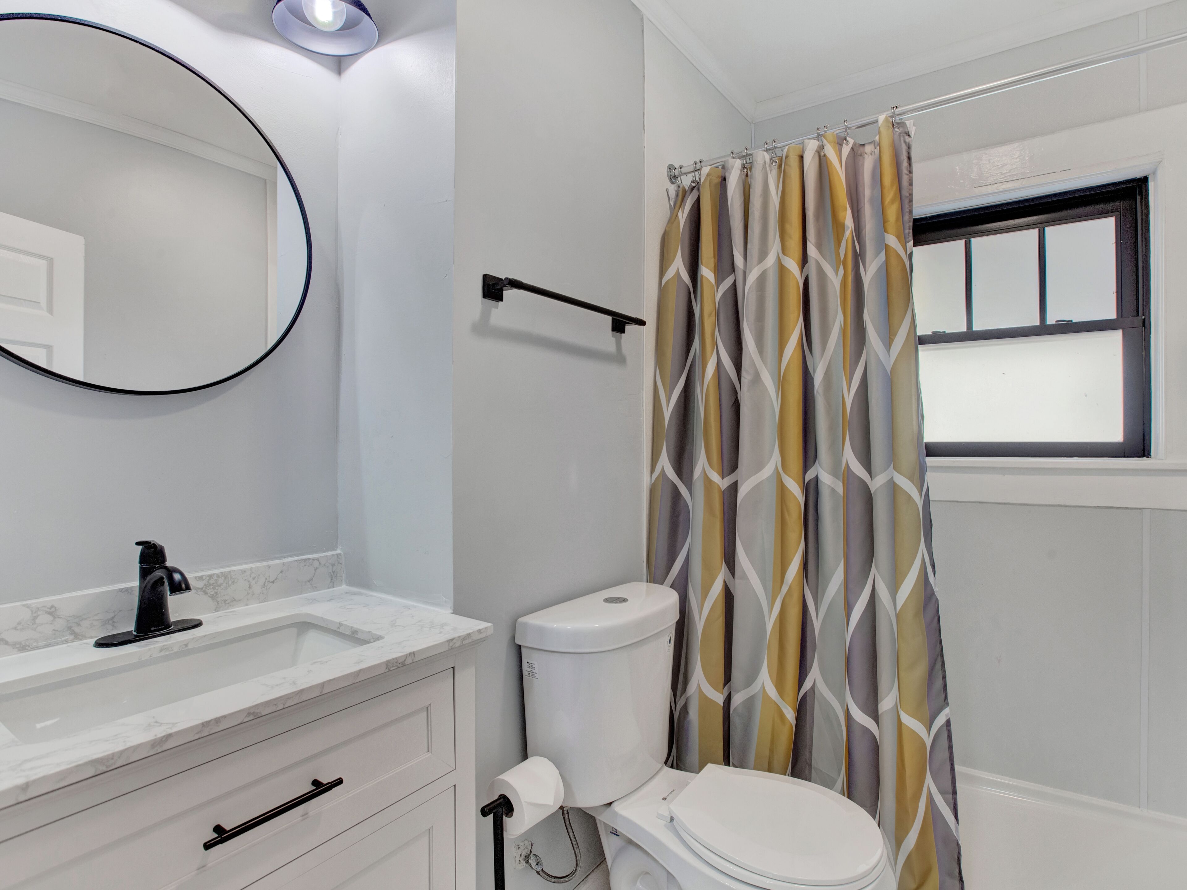 Un bagno con un water bianco, un lavandino con piano in marmo, uno specchio e una tenda della doccia con motivo geometrico.