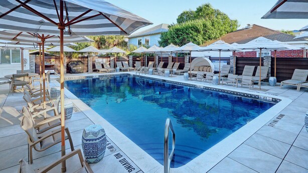 A swimming pool with lounge chairs and umbrellas, surrounded by a stone wall and a building in the background. - Entire Unit Steps from the Beach | Heated Pool Access (Tybee Island)