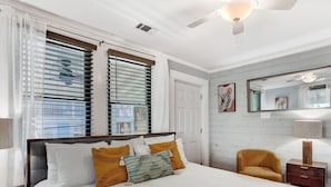A bedroom with a large bed, two windows with blinds, a ceiling fan, a mirror, and a small chair. - Entire Unit Steps from the Beach | Heated Pool Access (Tybee Island)
