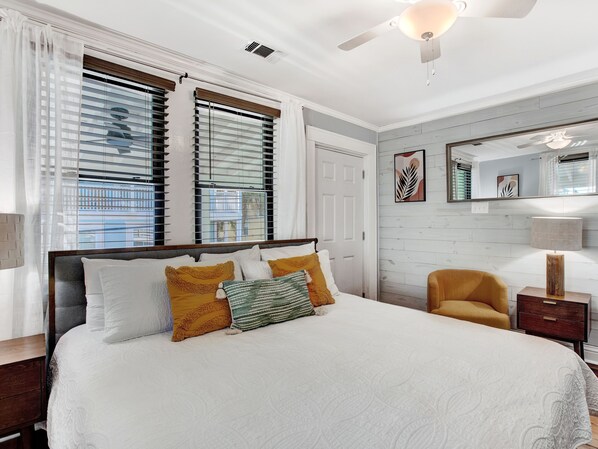 A bedroom with a large bed, two windows with blinds, a ceiling fan, a mirror, and a small chair.