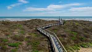 Un paseo marítimo de madera que lleva a un mirador con vista a una playa con poca vegetación y un cielo despejado.