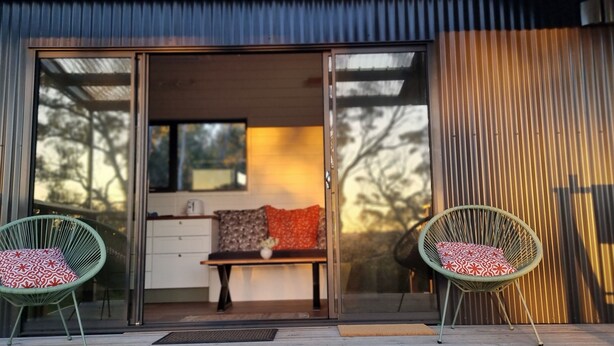 A modern building with a sliding glass door entrance, a bench with patterned cushions, and two wicker chairs with cushions. - Tiny House/Retreat in Adelaide Hills. Secluded, peaceful, country views. (Macclesfield)