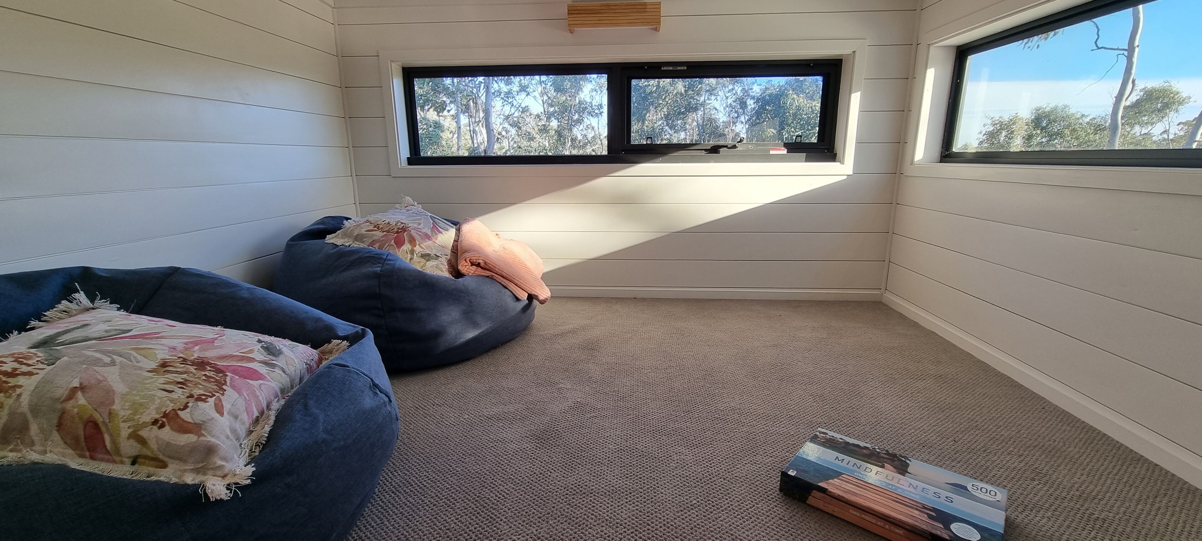 Une pièce avec un plafond haut, une grande fenêtre donnant sur des arbres, un fauteuil en forme de poire bleu, un autre, plus petit, et une boîte de livres posée sur le sol.