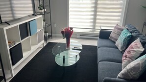 A modern living room with a glass coffee table, grey sofa, and a white entertainment unit with shelves. - Stylish Vacation Home (Dieppe)