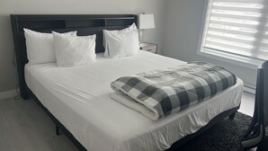 A neatly made bed with white bedding and a patterned throw blanket, a bedside table with a lamp, and a window with blinds. - Stylish Vacation Home (Dieppe)