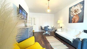 Un salon moderne avec un canapé jaune, un canapé bleu foncé, un coin repas avec une table et des chaises, un téléviseur fixé au mur et un tableau représentant des fleurs.