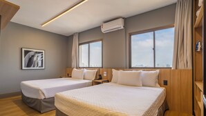 A hotel room with two beds, wooden flooring, large windows, an air conditioning unit, and a city view.