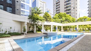 A swimming pool with tiled edges, surrounded by a paved walkway, and adjacent to a building with balconies and a covered seating area.