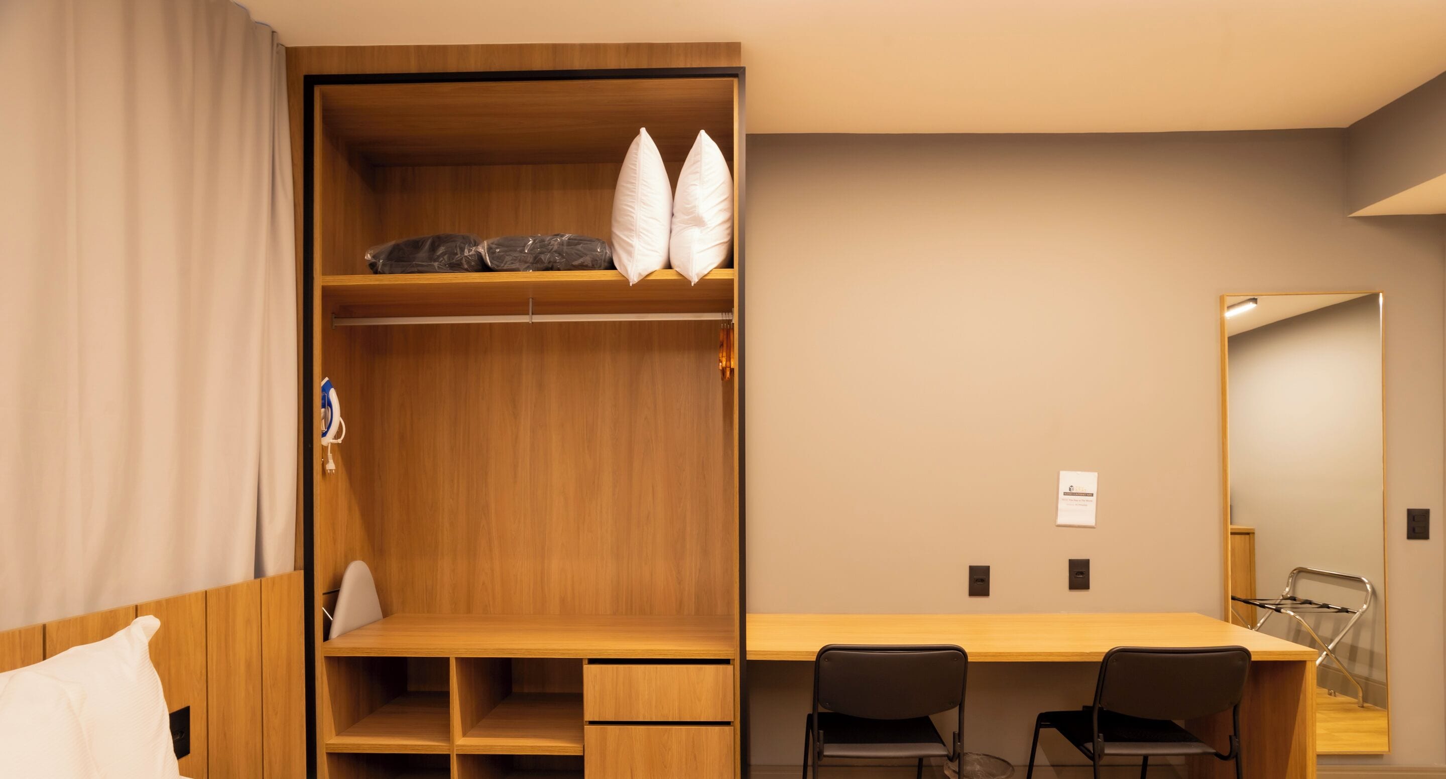 Une chambre d’hôtel moderne avec une armoire en bois, un bureau avec deux chaises et un miroir au mur.