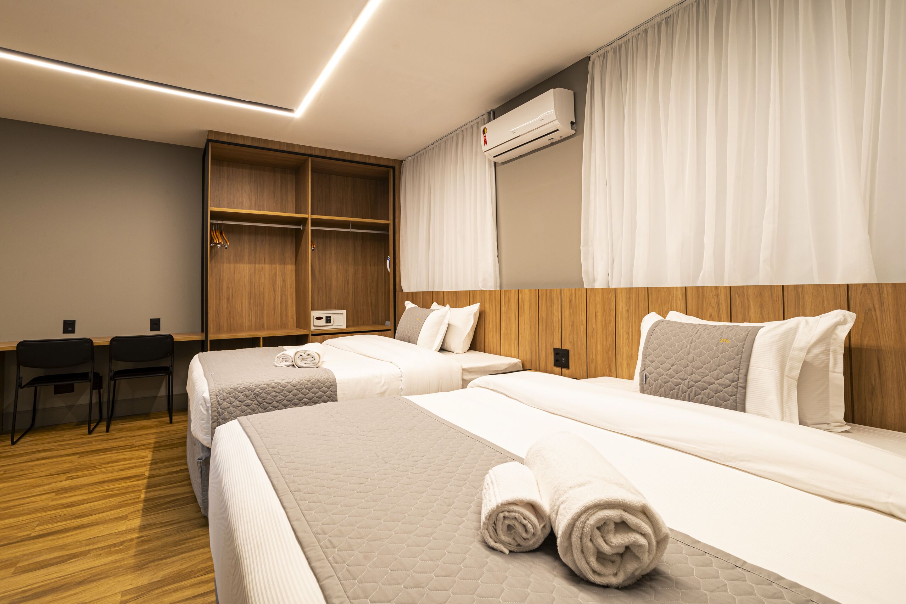 A hotel room with two beds, a wooden wardrobe, a desk with chairs, and an air conditioning unit.