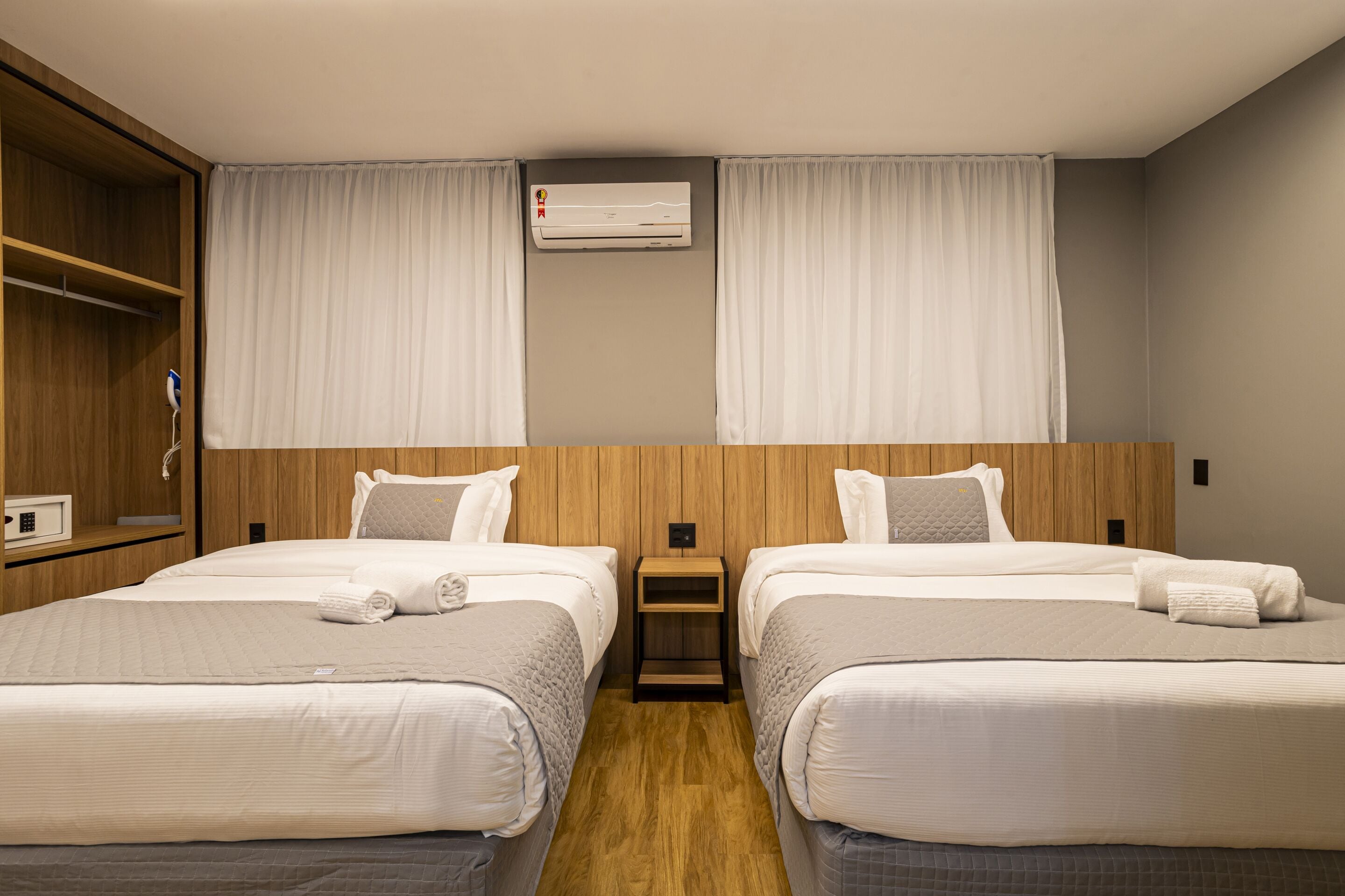 A hotel room with two beds, a wooden wardrobe, a nightstand, and an air conditioning unit.
