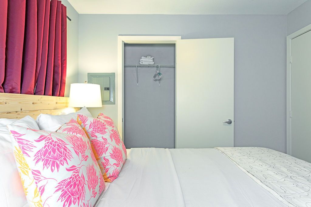 Un dormitorio con cama, almohadas, una mesita de noche con lámpara, una puerta con perchero para toallas y un espejo montado en la pared.