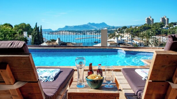 Ein Poolbereich mit Liegestühlen, einem Tisch mit Snacks, Getränken und Blick auf das Meer und die Berge.