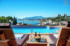 Un espace aménagé au bord de la piscine, avec des chaises longues, une table où sont servis des collations et des boissons, et une vue sur la mer et les montagnes.
