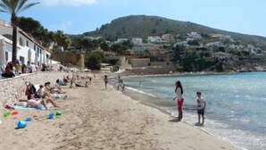 Ein Strand mit sonnenbadenden Menschen, spielenden Personen, einer Steinmauer und Gebäuden im Hintergrund.