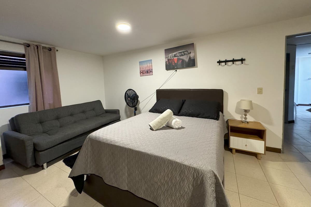 A bedroom with a bed, a grey sofa, a standing fan, a nightstand with a lamp, and a window with curtains.