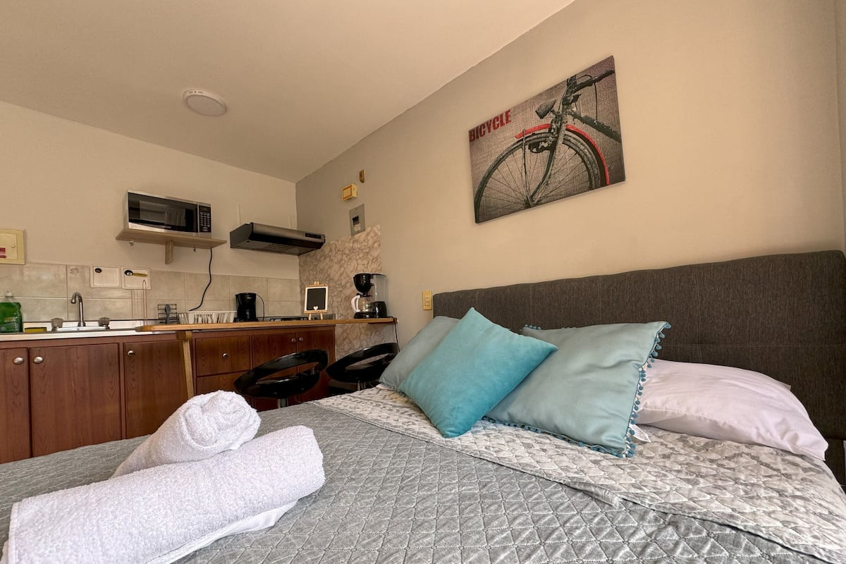 A hotel room with a bed, a headboard, a painting of a bicycle, a kitchenette, and a bathroom.