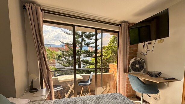 Une chambre avec un lit, une télévision, un bureau avec une chaise, un ventilateur et une vue sur l’extérieur.