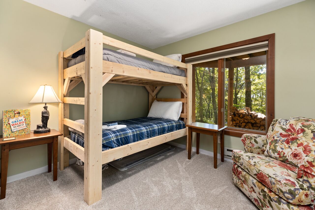 A bunk bed room with a wooden frame, a small table with a lamp, a floral sofa, and a window with a view of trees.
