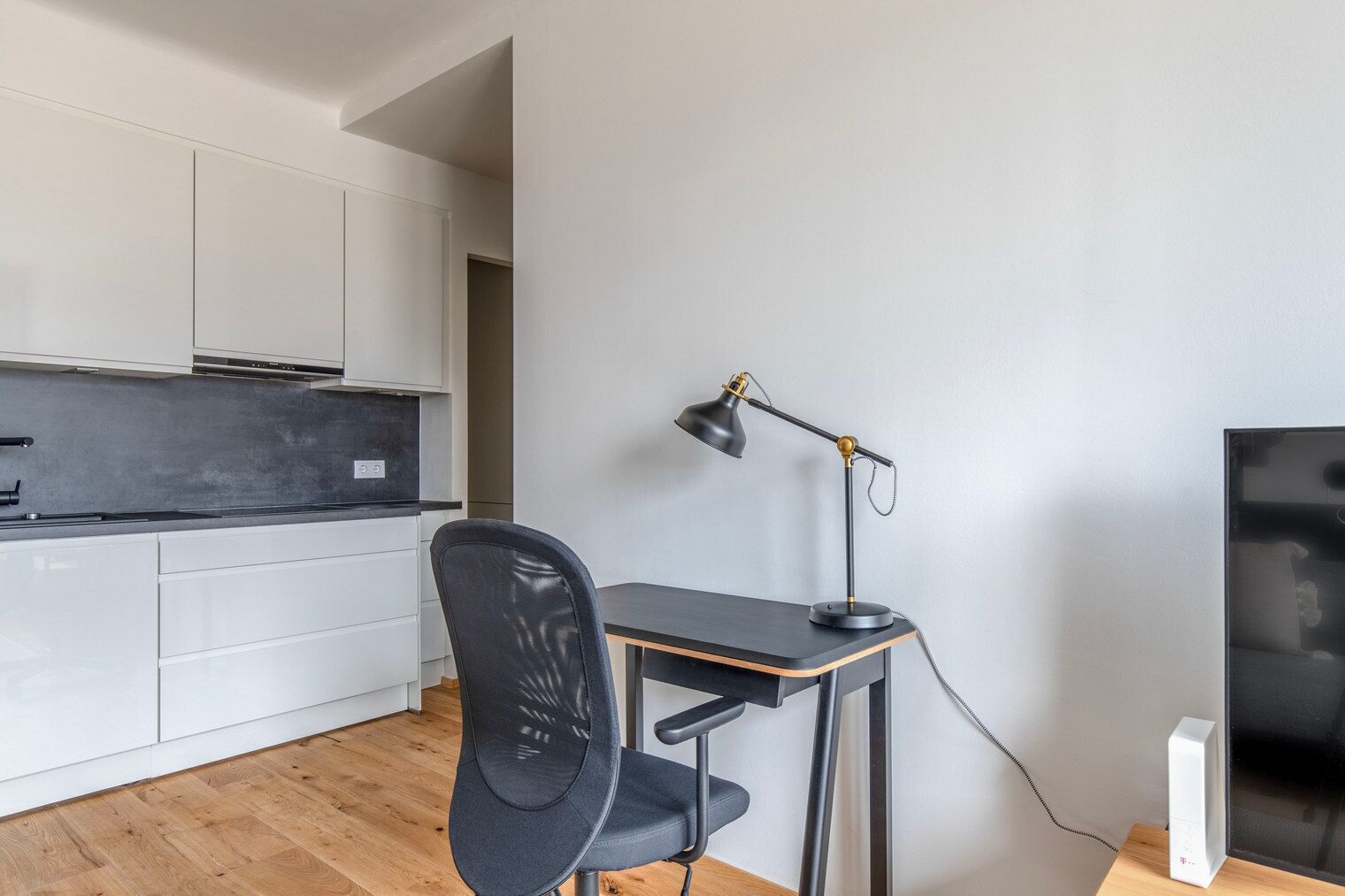 Cocina moderna con armarios blancos, encimera negra y suelo de madera. Se ve una silla de oficina negra, un escritorio y una lámpara de escritorio. Un televisor está sobre un mueble de madera a la derecha.