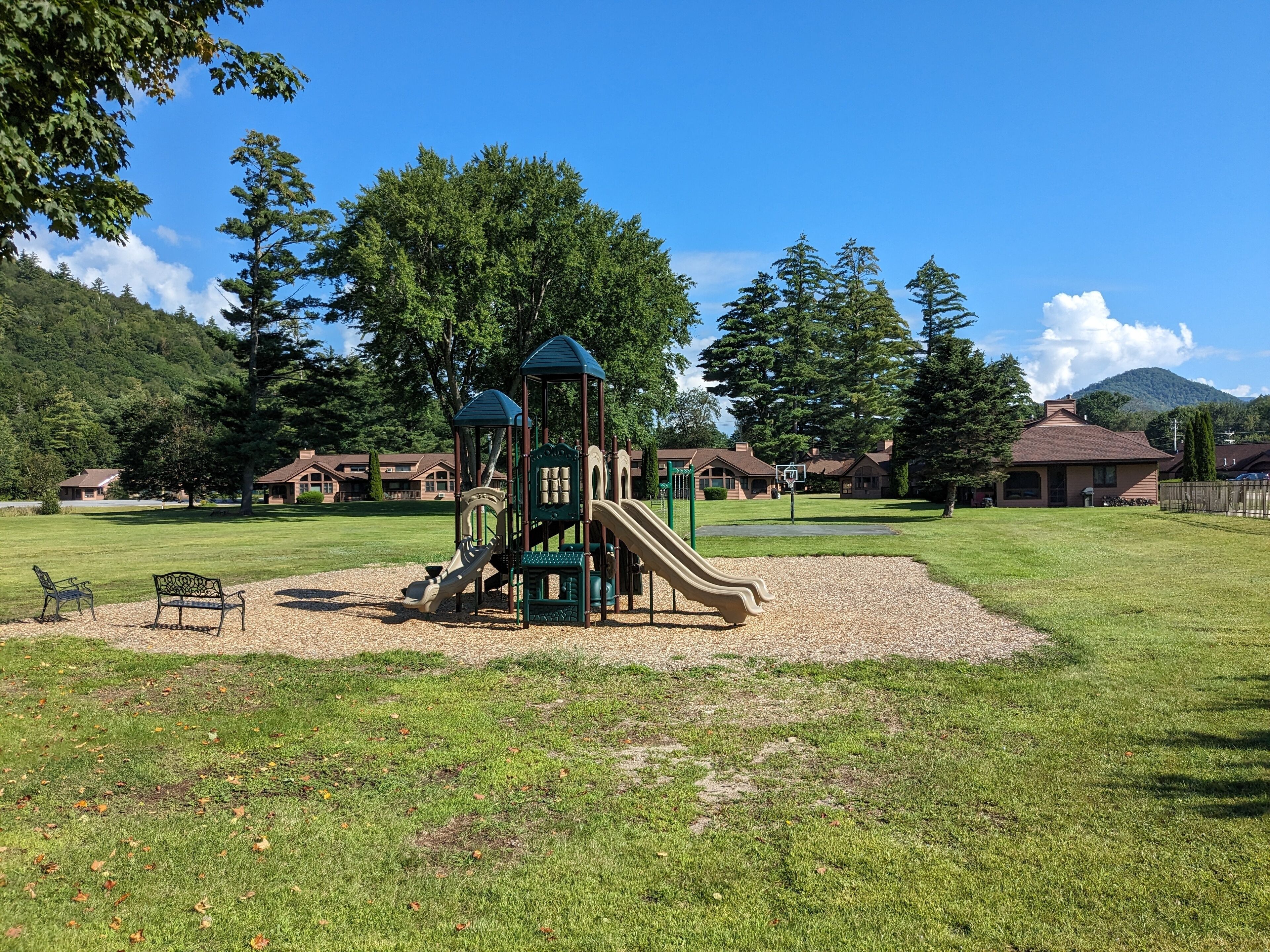 Un parque infantil con tobogán y estructuras para escalar, rodeado de césped y árboles, con edificios al fondo.