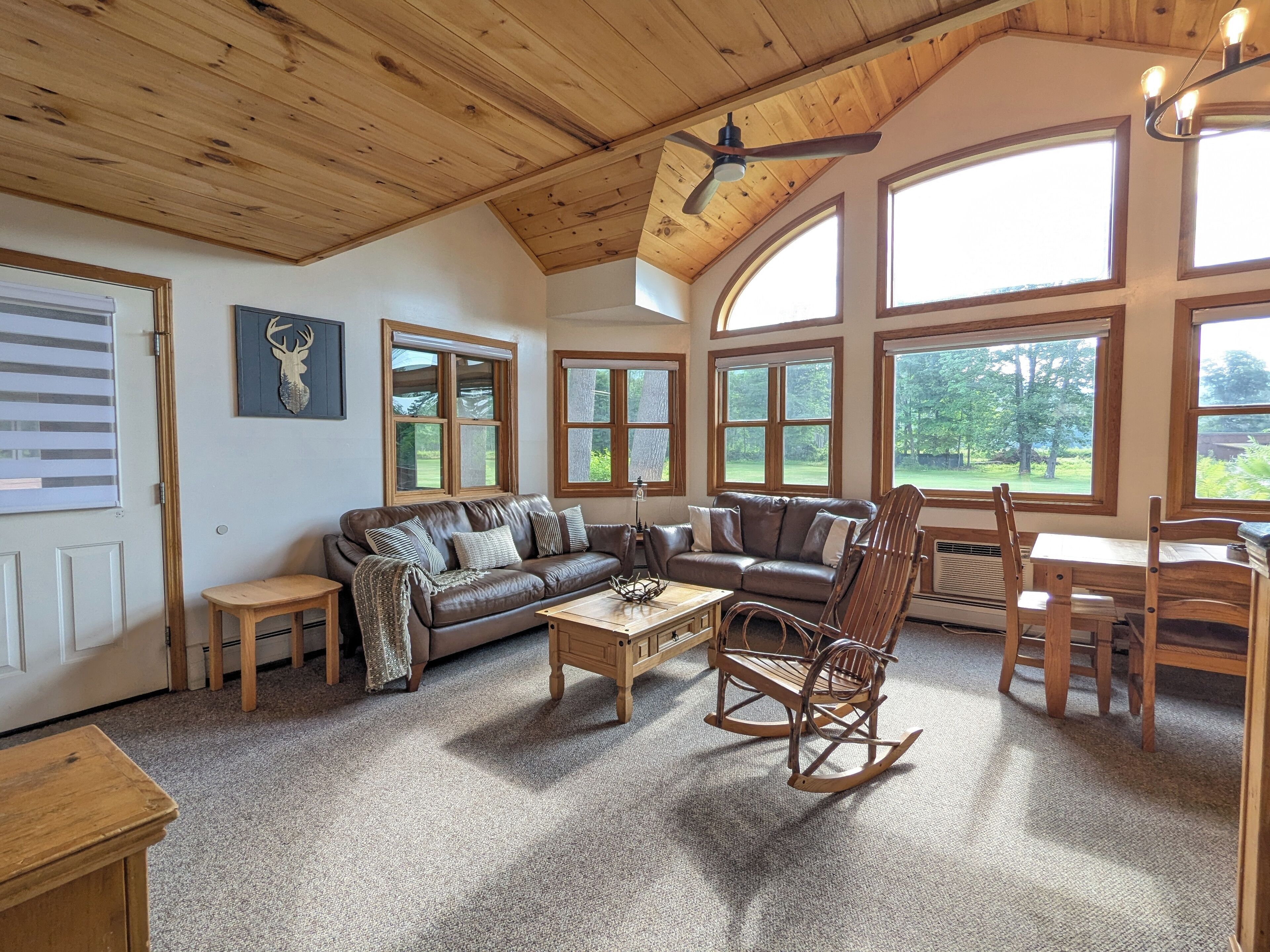 Ein gemütliches Wohnzimmer mit Holzbalkendecke, einem Ledersofa, einem Schaukelstuhl und großen Fenstern mit Blick ins Freie.