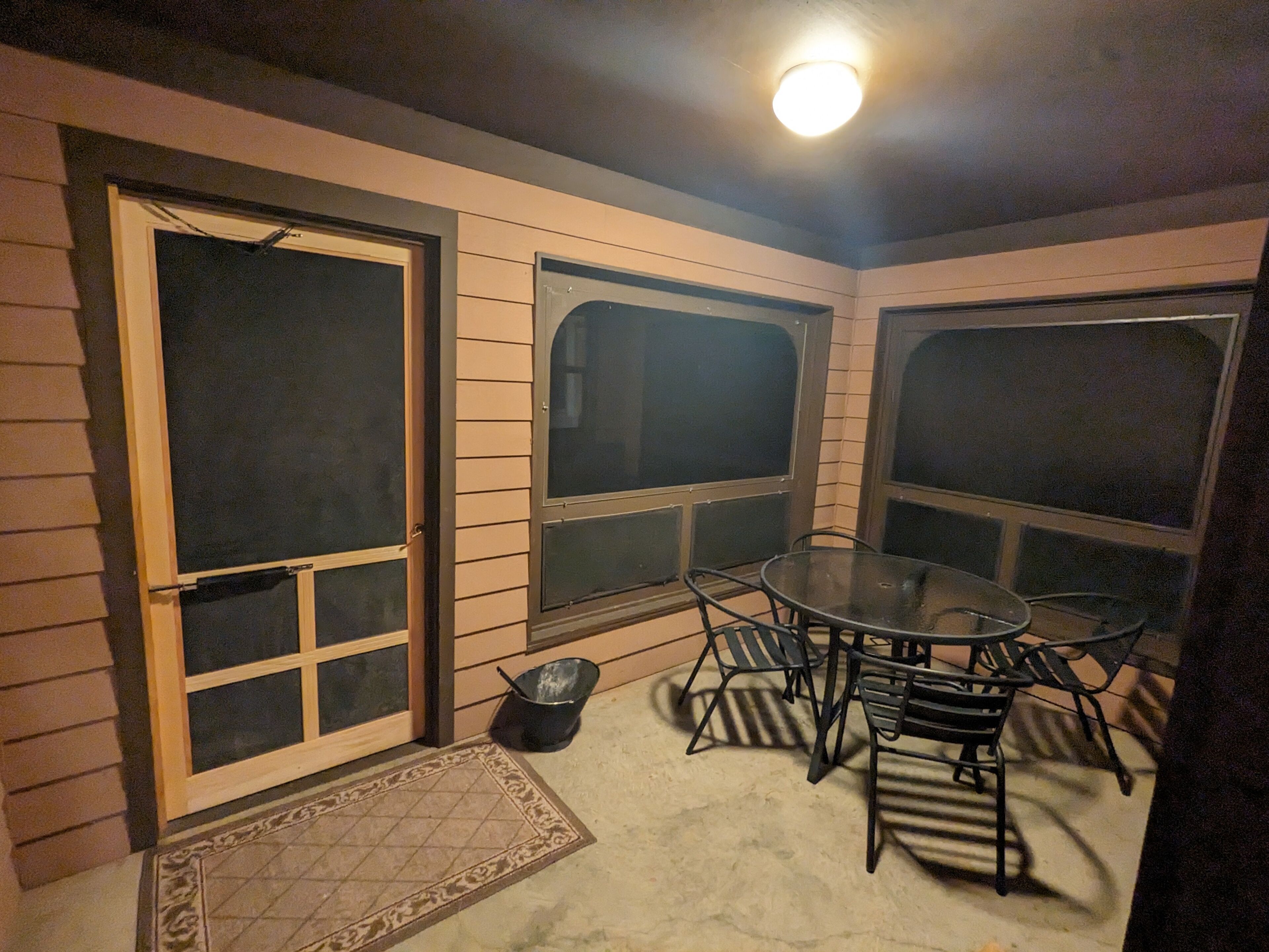 A covered patio area with a table and chairs, a small trash bin, and a door with a window.