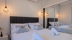 Un lit bien fait, avec du linge de lit blanc et des coussins à motifs noir et blanc, une table de chevet surmontée d’un vase et un grand miroir qui reflète la pièce.