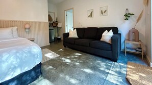 A bedroom with a bed, a dark-colored sofa, a small table, and a kitchen area in the background.