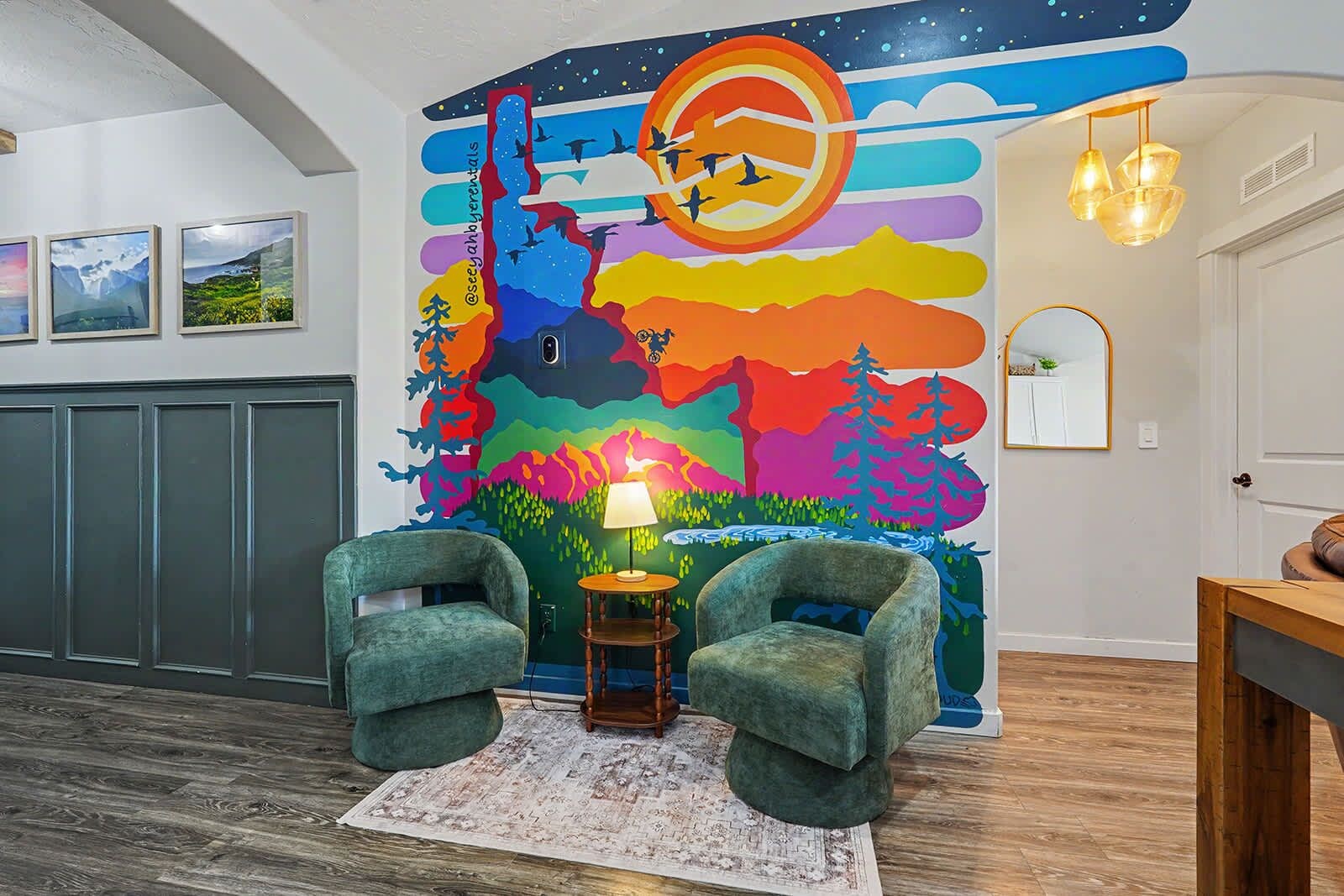 Une pièce décorée d’une fresque murale colorée, avec deux fauteuils verts, une petite table sur laquelle se trouve une lampe, et un plancher en bois.