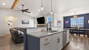 A modern kitchen with a central island, a dining area with a table and chairs, and a living room with a sofa and a ceiling fan.