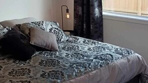 A bedroom with a bed, a window with curtains, a nightstand with a lamp, and a view of a wooden fence outside.