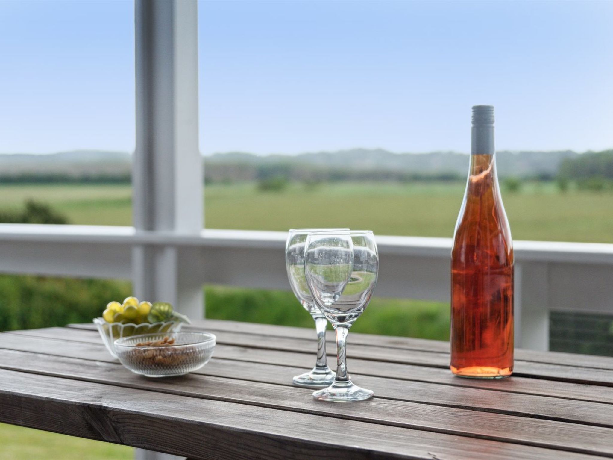 A wooden table with a bottle and two glasses on a balcony with a view of green fields.