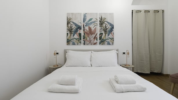 Een netjes opgemaakt bed met witte lakens en twee gevouwen handdoeken, een muur met drie schilderijen van tropische planten, en een gordijn voor het raam.