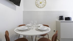 Een eethoek met een ronde tafel voor vier personen, compleet met borden, glazen en bestek. Daarboven hangt een klok aan de muur.