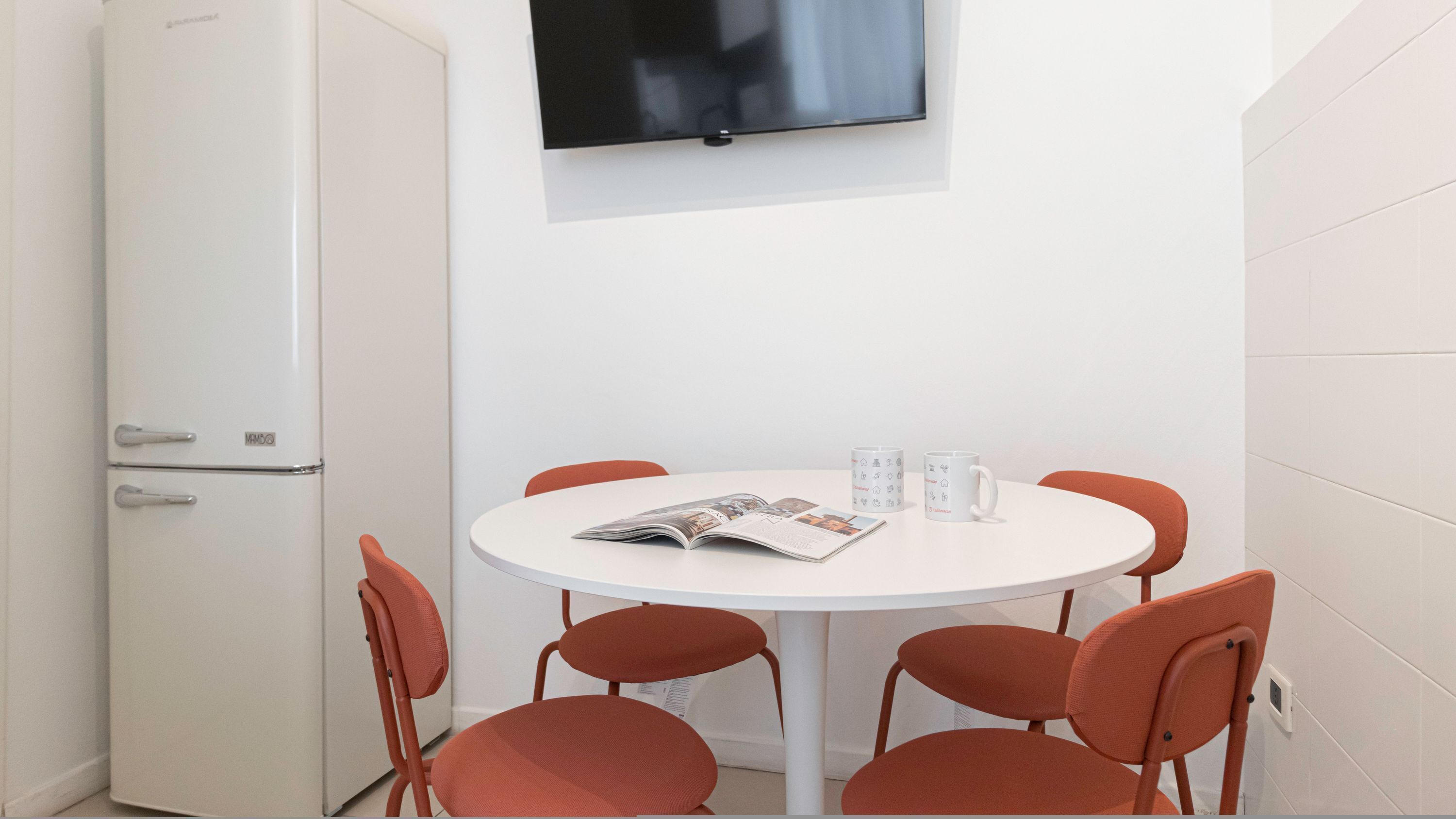 A small, modern room with a white table and four orange chairs, a white refrigerator, and a flat-screen TV mounted on the wall.