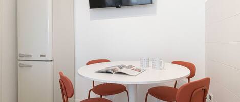 A small, modern room with a white table and four orange chairs, a white refrigerator, and a flat-screen TV mounted on the wall.