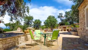 A stone patio with a glass table and chairs, surrounded by trees and a stone wall.