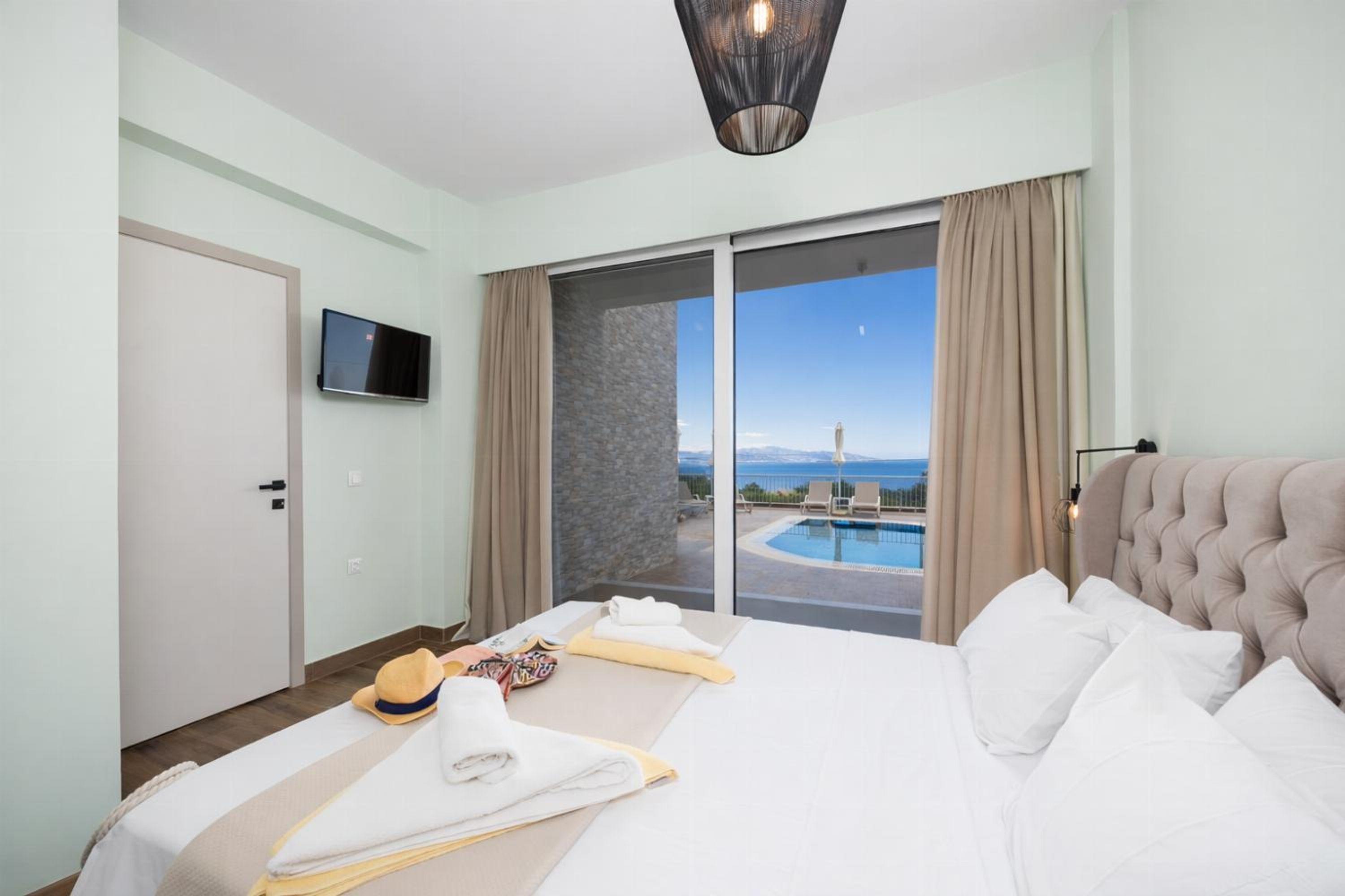 A hotel room with a bed, a TV, a view of a pool and the sea, and a sliding door to the outside.