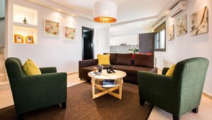 Ein modernes Wohnzimmer mit zwei grünen Sesseln, einem braunen Sofa und einem Couchtisch aus Holz. An den Wänden befinden sich Regale mit gerahmten Bildern und eine große, weiße Pendelleuchte.