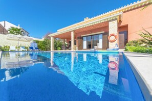 Una piscina de blau clar amb gandules i ombrel·les, una zona de seients coberta i un flotador a la paret.