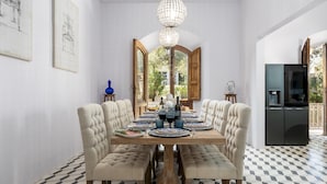 A dining room with a large wooden table set for six, surrounded by upholstered chairs. A chandelier hangs from the ceiling, and a large window provides a view of greenery outside. - Villa Silvia 10 Emma Villas (Terricciola)
