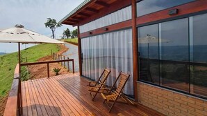 A wooden deck with two wooden chairs, a table, and a large window overlooking a landscape. - Chalé Sol no Horizonte  (Corrego do Bom Jesus)