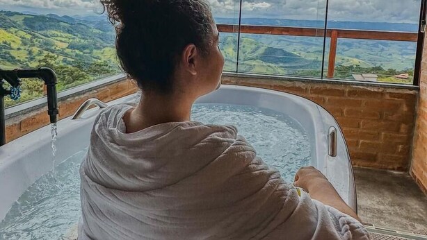 A person sitting in a bathtub with a view of a mountainous landscape through large windows. - Chalé Sol no Horizonte  (Corrego do Bom Jesus)