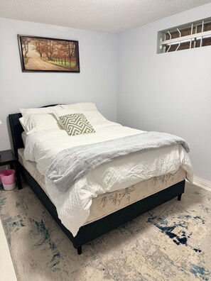 Un dormitorio con cama, mesita de noche, cubo de basura, un cuadro en la pared y una ventana con barrotes.