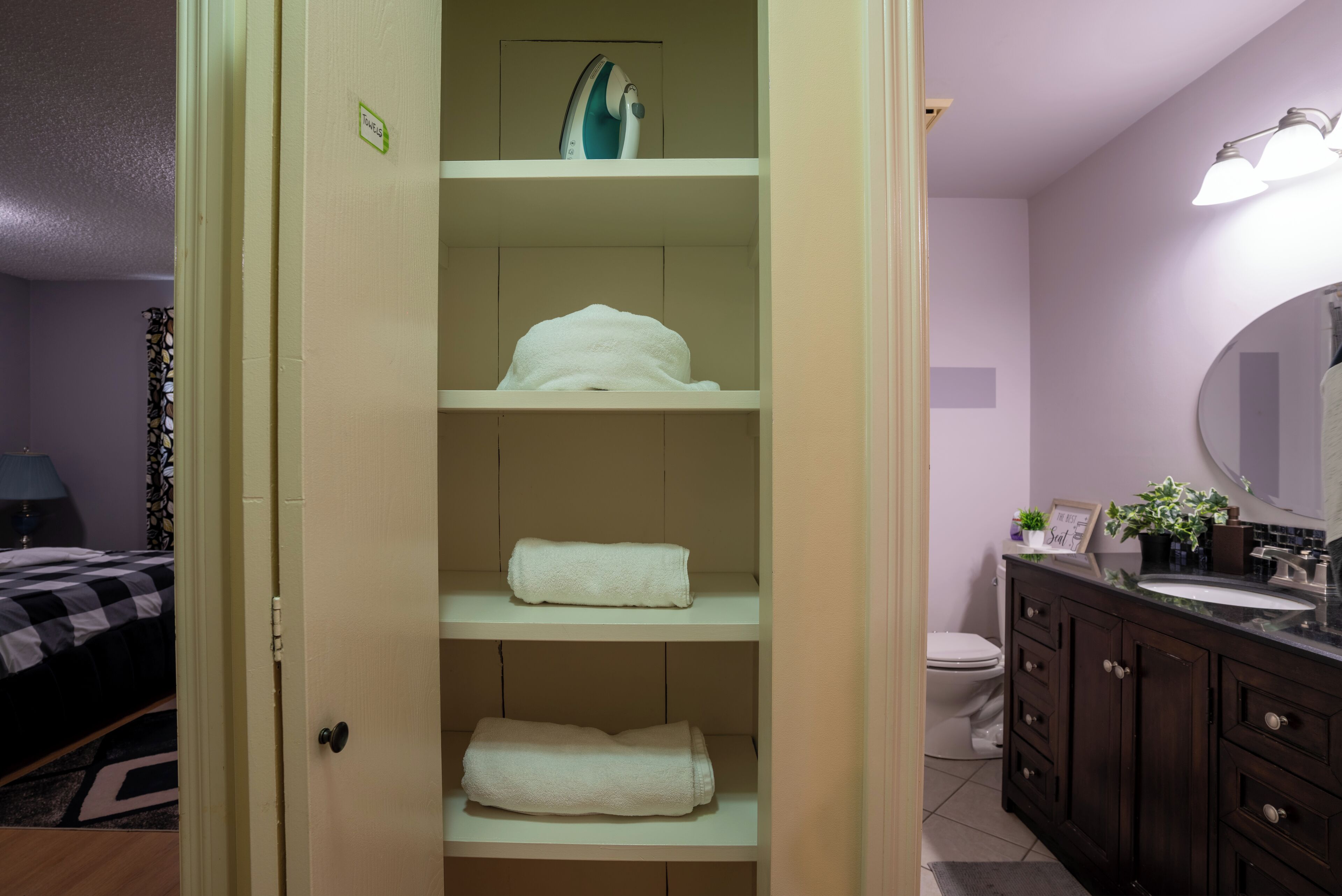 A bathroom with a toilet, sink, and vanity. There is a towel rack with folded towels and an ironing board on the wall.